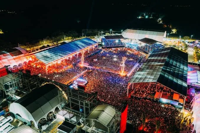 Vista aérea noturna de festival lotado com palcos e tendas iluminadas, filmagem com drone por PH Marx