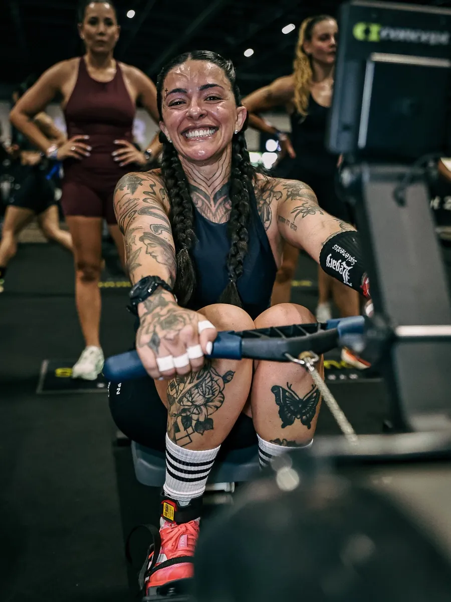 Atleta híbrida sorrindo na estação de remo do HYROX, foto de PH Marx