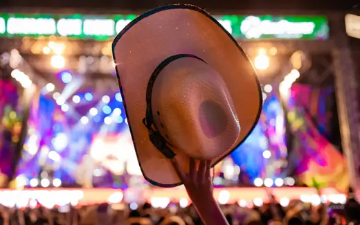 Chapéu de cowboy erguido diante de palco iluminado em show sertanejo, por PH Marx